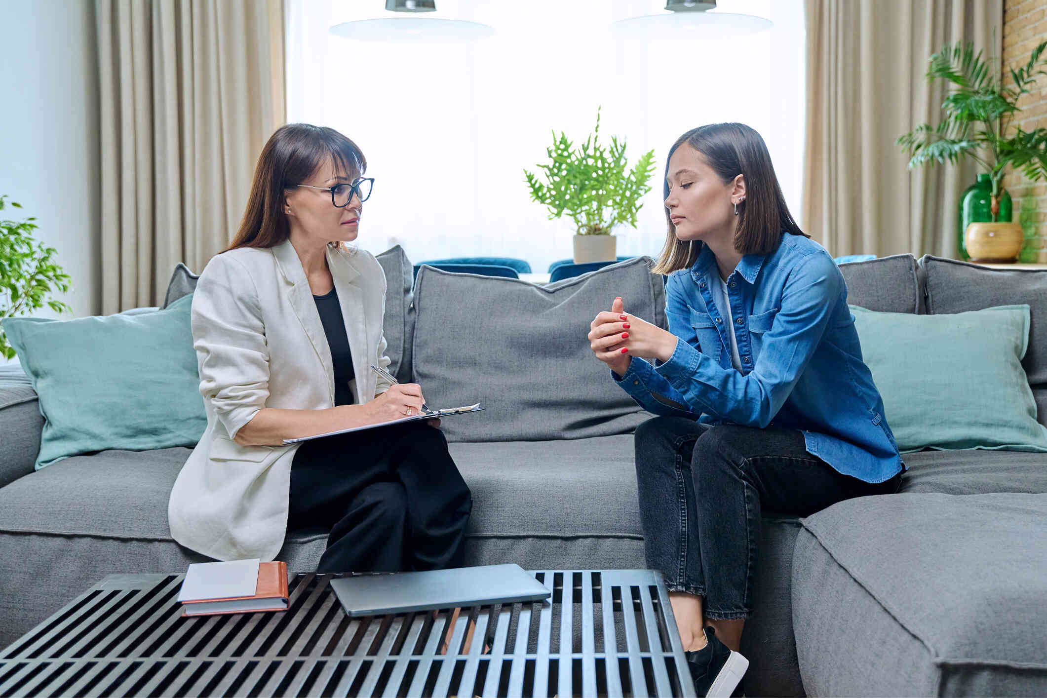 A woman ists hunched over on a couch while sitting next to her female therapist who is writting on a clipboard.